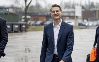 Een jonge man met bril en blond haar, gekleed in een blauw pak, loopt over een plein dat nat is van de regen. Op de achtergrond staan gebouwen en auto’s.