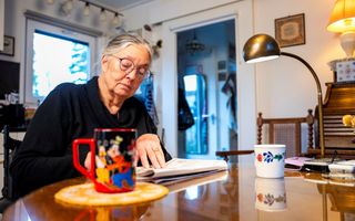 Een oudere vrouw met bril zit aan een tafel in een woonkamer, met een dubbelgevouwen krant voor zich. Op tafel staan twee mokken