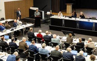 Zaal vol mensen die achter rijen tafels zitten, van bovenaf gezien. Links voorin staat een vrouw achter een katheder.