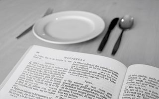 Een Bijbel die ligt opengeslagen bij Mattheüs 6 (over het vasten). Ook zijn een leeg bord en bestek zichtbaar.