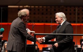 Twee donker geklede mannen schudden elkaar de hand op het podium in een concertzaal. De man links houdt een beeldje in zijn andere hand.