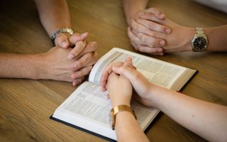 Drie paar gevouwen handen liggen op tafel naast een opengeslagen Bijbel.