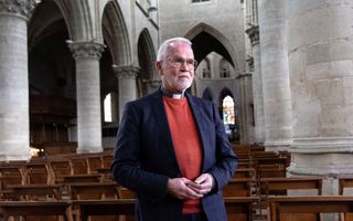 Man met grijs haar en grijze baard, blauwe jas en oranje trui tussen de pilaren van een monumentale kerk.