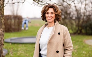 Vrouw van middelbare leeftijd met donker haar en beige jas in een tuin.