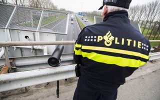 Politieman met pet kijkt naar een camera op een statief op een viaduct. Onder hem rijden auto’s over de snelweg.  