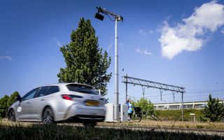 Een grijze auto rijdt langs een hoge paal waar een camera aan hangt. Op de achtergrond fietst een vrouw met een blauw jasje aan.