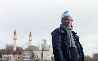Een man met een jas, een sjaal en een hoofddeksel kijkt schuin opzij. Achter hem staat een moskee met twee minaretten.