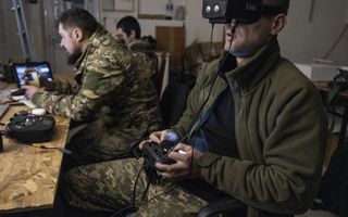Een man in camouflage-uniform zit met een VR-bril op zijn hoofd en een afstandsbediening in zijn handen achter een tafel. Achter hem zitten nog twee militairen.