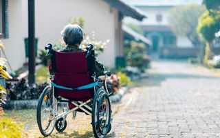Een vrouw zit in een rollator met op de achtergrond een riante villa. 