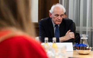 Man in donker pak met blauwe stropdas, zittend aan een tafel.