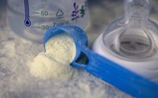 Een blauw schepje met wit poeder ligt naast een leeg drinkflesje voor baby’s.