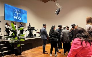 Mensen staan in een kerk rond een man met zwart haar, gekleed in een lichtblauw kostuum; op een podium staat een band.