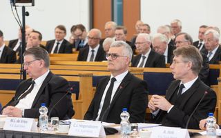 Drie mannen in zwarte pakken achter een vergadertafel.