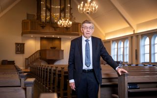 Een man in een blauw pak staat naast donkerbruine kerkbanken. Op de achtergrond de preekstoel en daarboven het orgel.