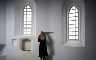 Een vrouw in een zwarte jurk staat met het gezicht naar een witte kerkmuur met aan beide kanten hoge ramen.