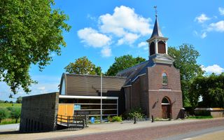 Een klein kerkje, met een wit torentje, aan de weg. Erachter is grasland zichtbaar.