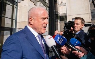 Een bijna kale man in een blauw pak spreekt in enkele microfoons van journalisten die hem bevragen en filmen voor een gebouw.