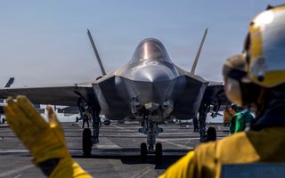 Man met helm gebaart naar gevechtsvliegtuig aan boord van vliegdekschip.