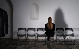 Een vrouw in het zwart in de kerk op een stoel die naar de muur gericht is, met naast haar vijf lege stoelen die andersom staan. Links van haar is een deel van een zwart colbert zichtbaar.