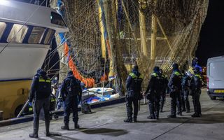 Politieagenten staan in het donker op een kade, waar een viskotter ligt aangemeerd. Netten hangen aan gele masten van het schip.