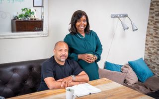 Een echtpaar met donkere huidskleur. De man heeft een gemillimeterd kapsel, baardje en een zwart T-shirt, de vrouw een petrolkleurige jurk en donker, loshangend haar. Op tafel ligt een geopende Bijbel en staat een koffiekopje.