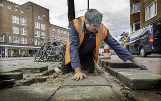 Een oudere stratenmaker met een oranje hesje is in de weer met zware stoeptegels. Op de achtergrond staan gebouwen.