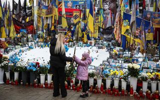 Een vrouw en een meisje leggen bloemen op een plein waarop bloemen, vlaggen en kaarsjes staan.