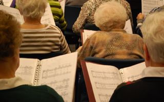 Foto van oudere vrouwen in kooropstelling, met bladmuziek in de handen. De foto is van achteren genomen.