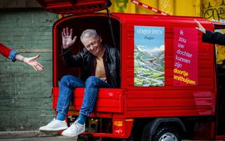 Foto van een oudere man in spijkerbroek. Hij zit in de laadbak van een kleine rode auto en zwaait met zijn rechterhand.