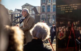 Een man in een bruine jas spreekt voor publiek dat wordt weerspiegeld in een zwarte zuil.
