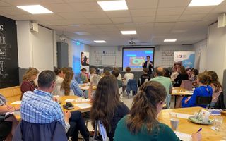 Een zaal vol mensen die aan tafeltjes zitten. Voorin een spreker, in pak.