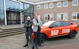 Twee mannen staan met de armen over elkaar bij een knaloranje auto met het logo van de SGP. Op een zijdeur staat ook een verkiezingsslogan: koersvast & betrokken.