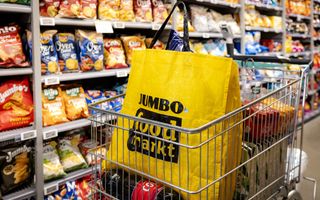 Een winkelwagen waarin een Jumbotas staat. Die is geel. Het logo staat in een zwart vak. Op de achtergrond zie je schappen met onder andere chips.