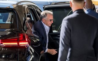 Een man in donker pak, met een zonnebril op, stapt uit een auto.