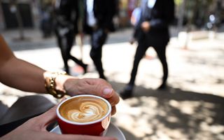 Foto van een kop koffie die door twee vrouwenhanden wordt vastgehouden. Op de achtergrond lopen mannen in pakken voorbij.