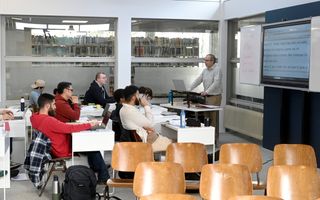 Studenten zitten in collegebankjes. Vooraan de docent, met naast hem een digibord waarop de Griekse tekst van de eerste Johannesbrief te zien is.