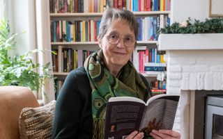 Vrouw met kort grijs haar, bril en een groene sjaal zit met een boek op de bank en kijkt je glimlachend aan.