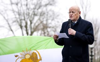 Een kale man met een montuurloze bril staat met een microfoon en een A4’tje in zijn handen voor een Iraanse vlag.