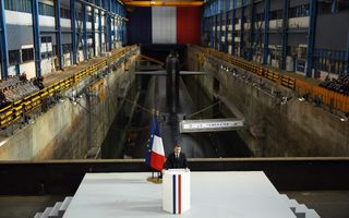 Een man in pak met stropdas staat achter een spreekgestoelte voor een dok waarin een onderzeeër ligt.