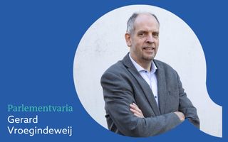 Foto van een glimlachende en wat oudere, kalende man in blauwgrijs colbert, zonder stropdas die armen over elkaar heeft.