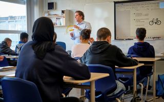 Een docent met een lichtblauw overhemd en bruine krullen houdt een grote geodriehoek in zijn hand. Op een digibord staat een wiskundeopgave. Leerlingen kijken naar de docent.