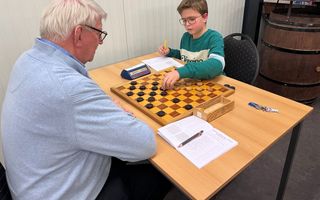 Tafel met daarop dambord. Tegenover elkaar zitten een tiener en een zeventiger.