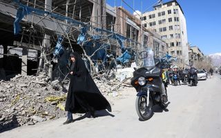 Een vrouw in een zwart gewaad loopt langs een beschadigd gebouw, waarvan een berg brokstukken op straat ligt.