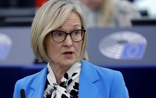 Vrouw van in de zestig met kort blond haar, zwarte bril en felblauw jasje spreekt.