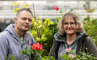 Een man en vrouw staan in een kas tussen groene planten en een rode geranium op de voorgrond. 