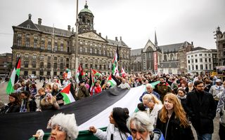Tegen een achtergrond van statige gebouwen staat een groep mensen met zwart-wit-groen-rode vlaggen.