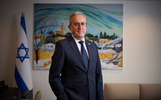 Man met bril in donker pak staat voor schilderij en blauw-witte vlag.