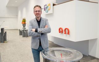 Een man met een baardje en een bril staat met de armen over elkaar bij een doopvont. Achter hem staat een witte preekstoel. 