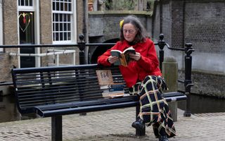 Vrouw in rode jas en met geblokte broek leest een boek op een bankje buiten in een stad. Naast haar op het bankje ligt een stapel boeken.