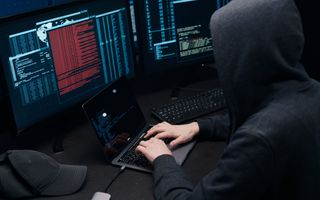 Een man met een capuchon op zit achter een laptop. Voor hem schermen met tal van data.